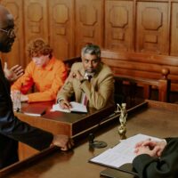 Man in a blue suit speaks at a podium in a courtroom as others at the table sign documents in the background?courtroom scene with a presenter at the lectern and two attorneys reviewing paperwork.